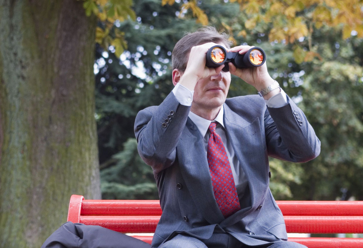 Businessman on a red park bench looking through binoculars.