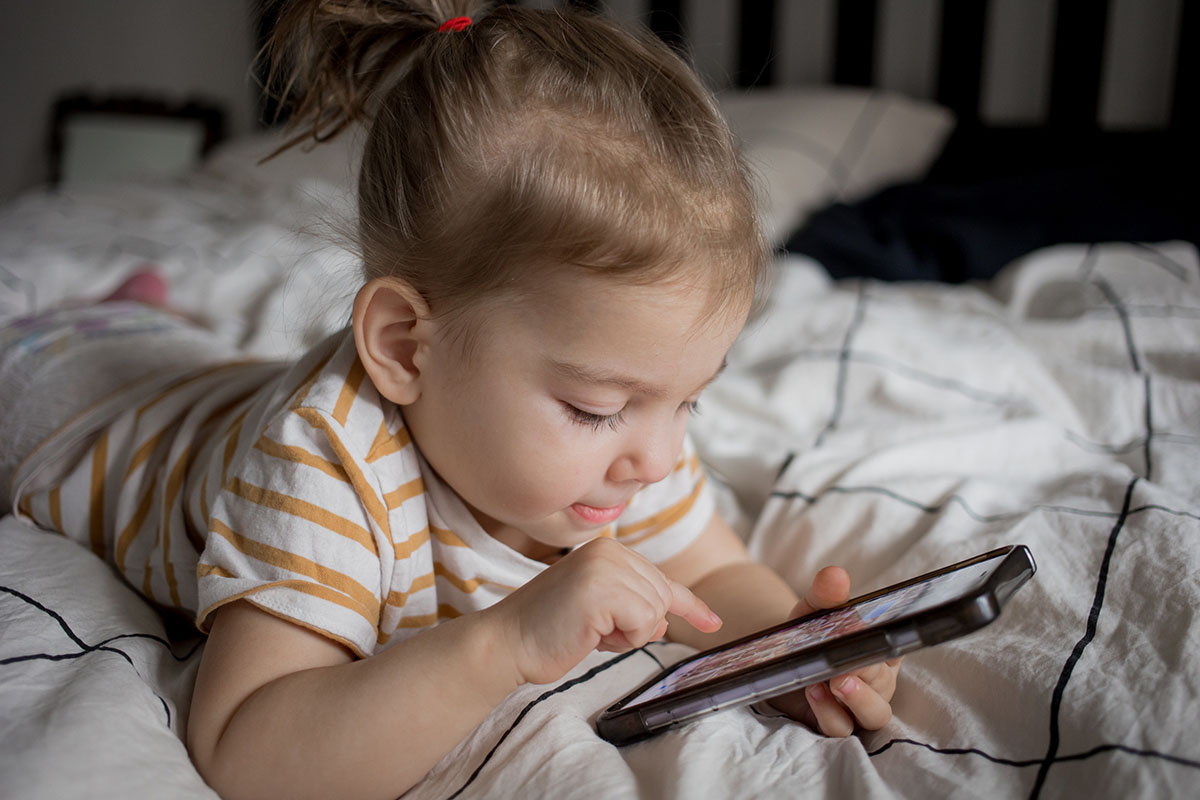 Little toddler aged girl playing with smartphone laying on the bed.