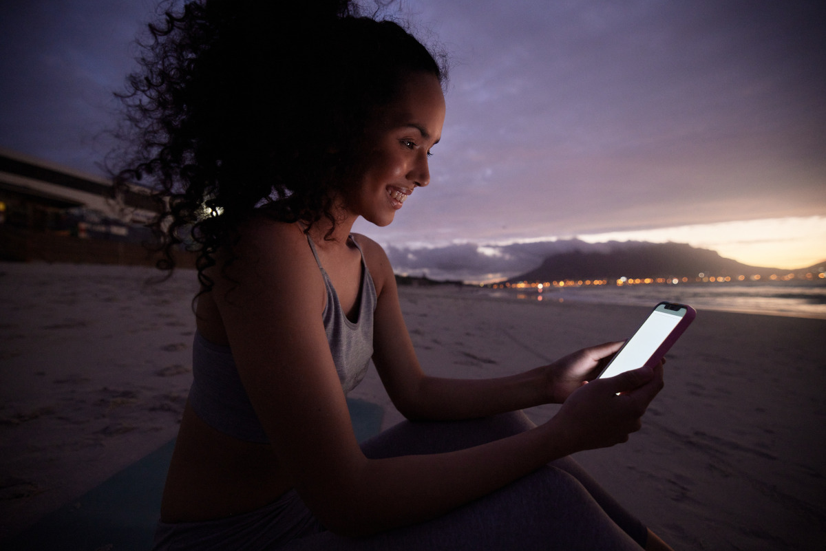 Fit young woman using her cellphone while sitting on the beach at night.